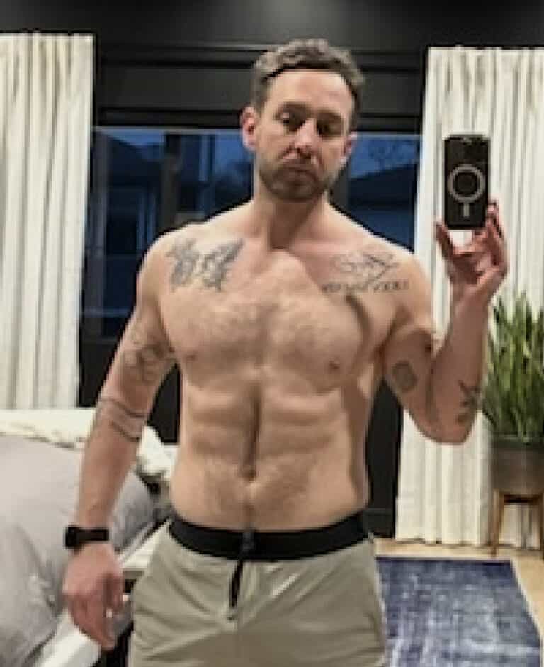 A shirtless man with tattoos, possibly an online fitness coach, takes a mirror selfie in a bedroom with curtains, a bed, and a plant visible in the background.